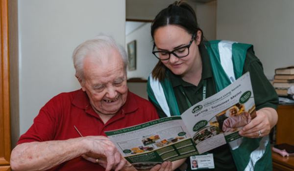 A young woman with a lanyard and hi-vis jacket shows an elderly man a menu. He is smiling and pointing to an item on the menu.