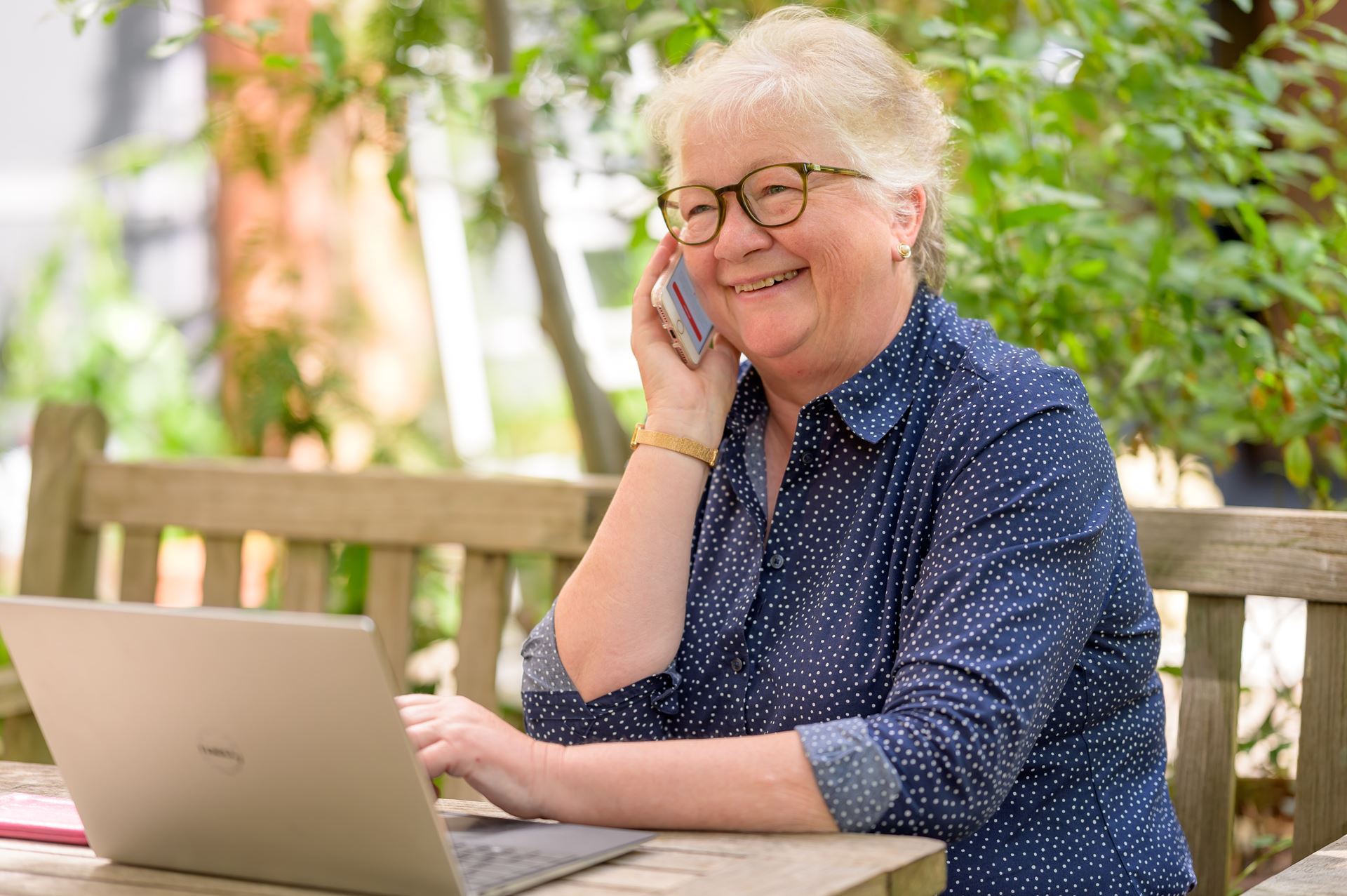 A woman with grey hair and glasses is speaking on the phone. She is smiling.